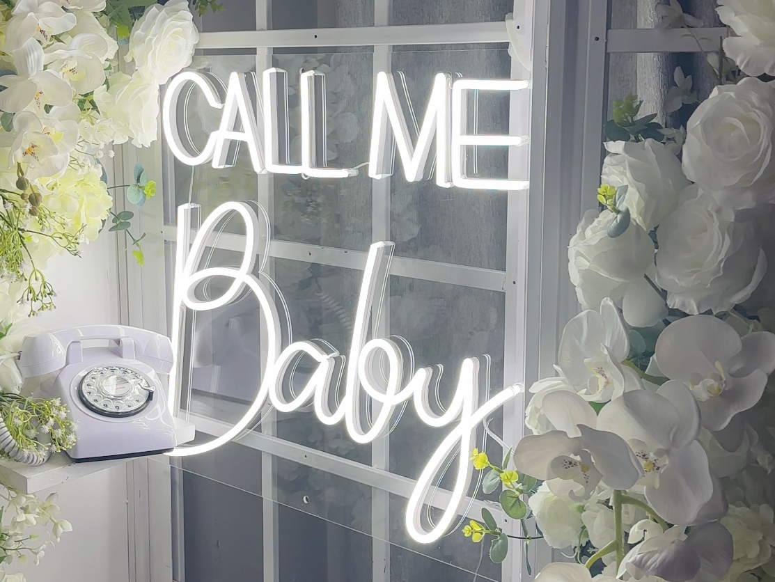 A "call me baby" neon light surrounded by flowers and a vintage telephone 